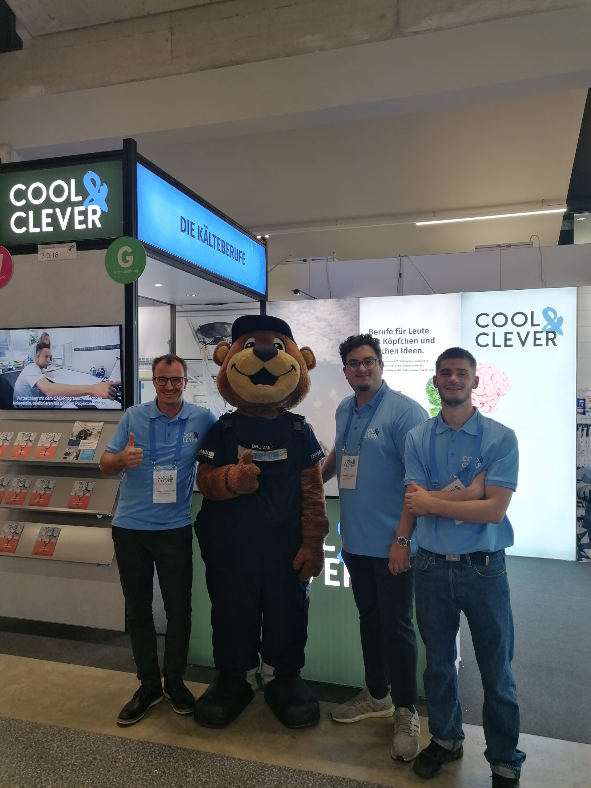 Berufsbildner Roger, Maskottchen Brummi, Georgios und Noe stehen am Stand der Kältetechnik, Cool & Clever. Die Lehrlinge haben die Lehrabschlussprüfung erfolgreich bestanden.